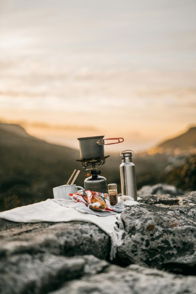 Olla de camping y termo de acero inoxidable colocados sobre una roca al atardecer