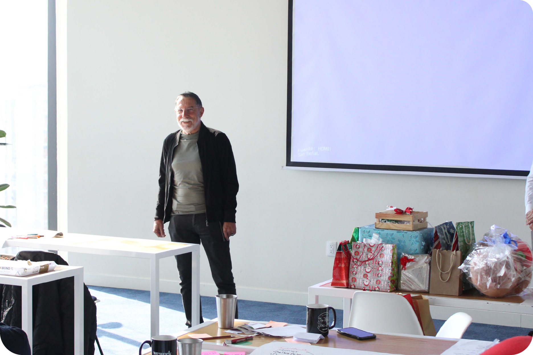 Hombre adulto sonriendo de pie en una oficina frente a una pantalla de proyección blanca, junto a una mesa llena de regalos envueltos y bolsas festivas.