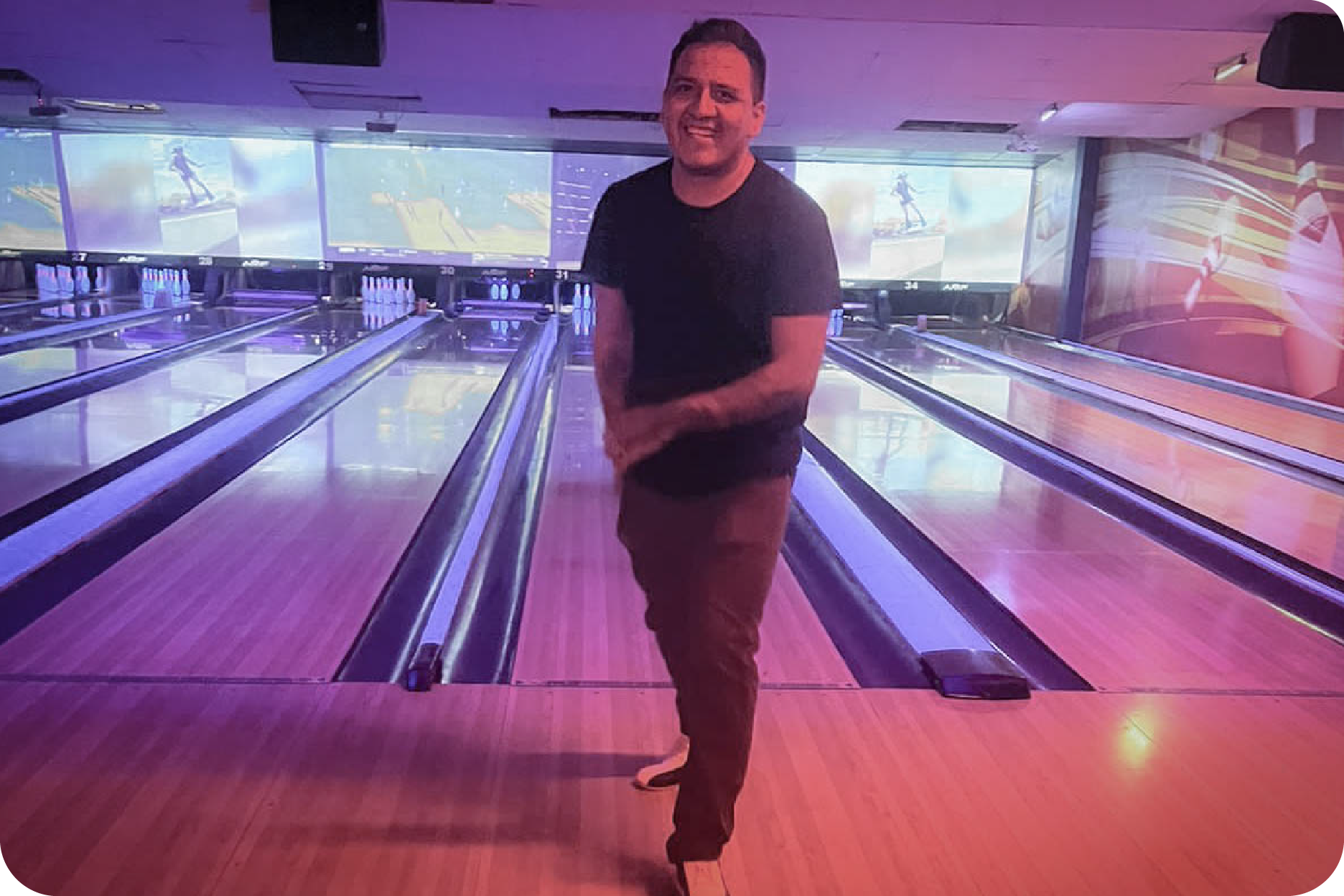 Hombre sonriendo de pie en medio de una pista de boliche (bowling) con iluminación neón púrpura y pantallas al fondo.