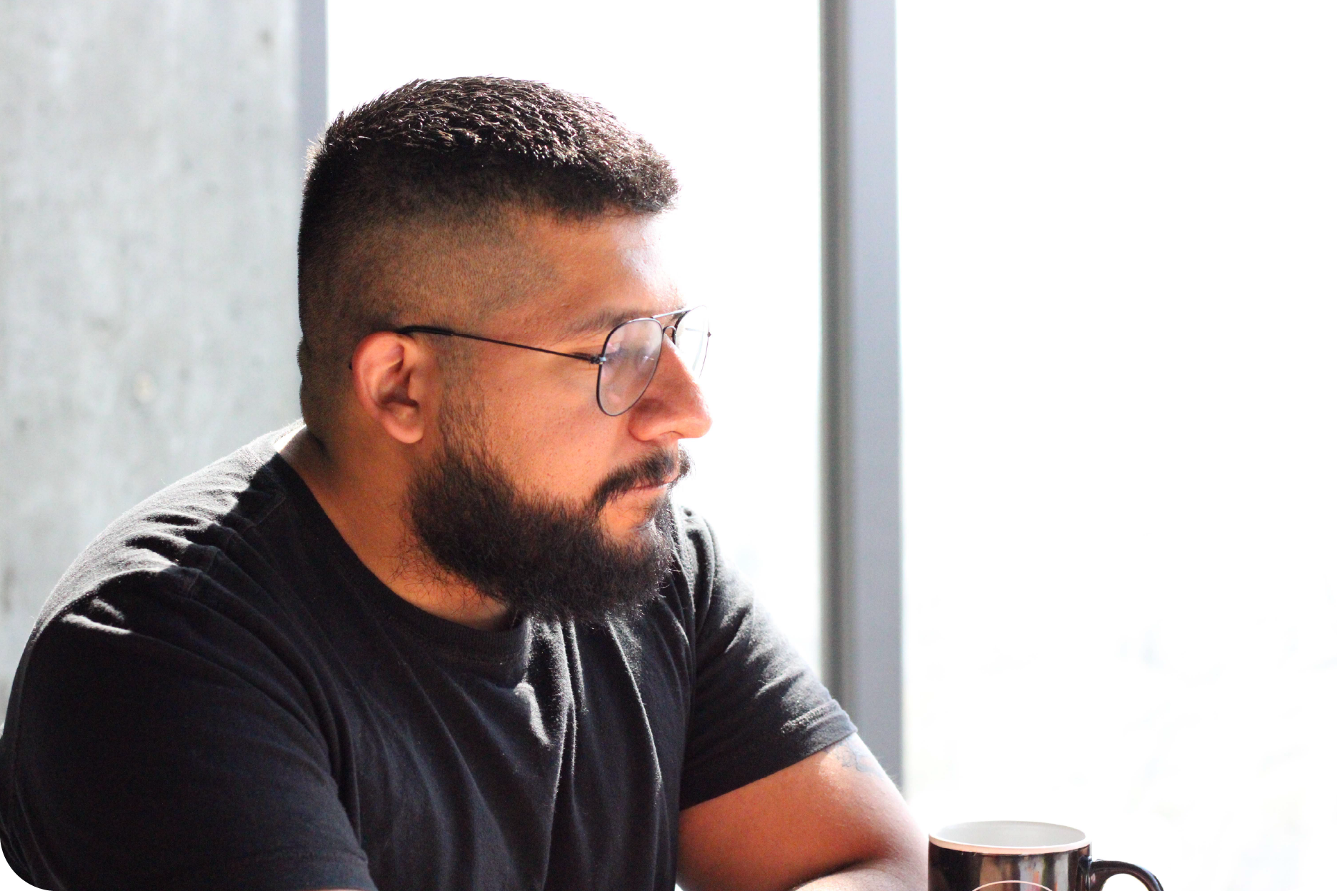 Primer plano de perfil de un hombre con barba y lentes, vistiendo una camiseta negra y mirando pensativo hacia una ventana con una taza de café enfrente.